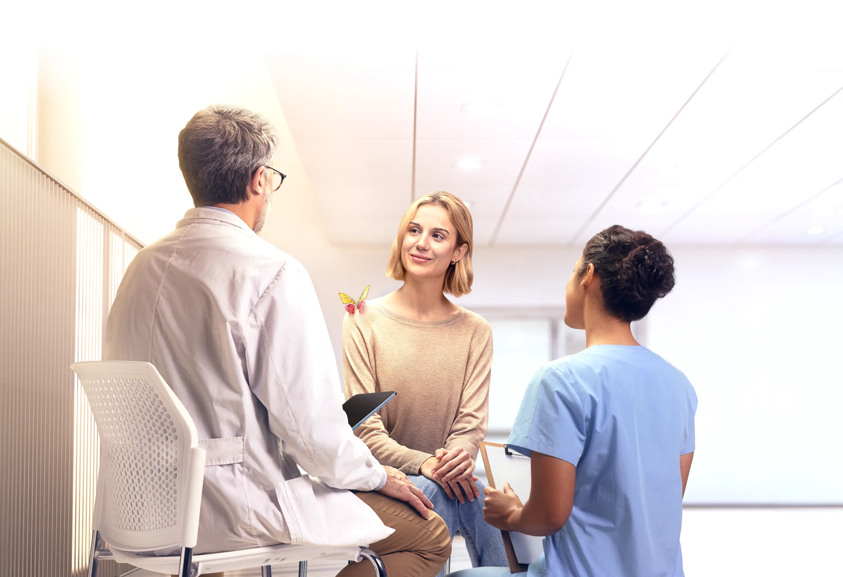 Photo image of an adult female patient speaking with doctor and nurse in a hospital setting, with an engineered mechanical yellow and magenta butterfly on her shoulder